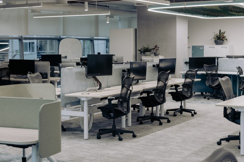 The image depicts a modern, open-plan office with a clean and organized layout. The workspace features multiple desks equipped with computers and ergonomic chairs. The desks are separated by low dividers, creating individual workstations while maintaining an open and collaborative environment. Overhead lighting and large windows provide ample illumination, enhancing the bright and airy atmosphere of the office. The decor includes minimalistic elements with a few potted plants adding a touch of greenery. The overall design emphasizes functionality and comfort, making it a conducive environment
